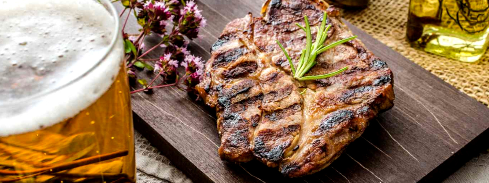 A imagem mostra um steak em uma tábua de corte ao lado de um copo de cerveja na carne