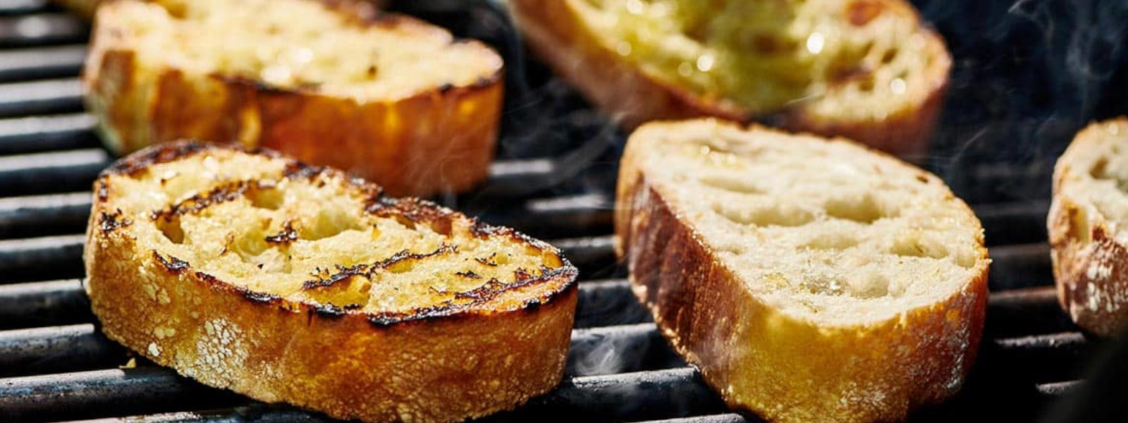 A imagem mostra uma grelha com pães no churrasco sendo preparados
